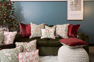 A green velvet sofa with a Red Blocking Velvet Pillow sits before a blue wall, flanked by a Christmas tree and geometric art. A knitted pouf and plush rug add to the cozy, holiday-inspired space.