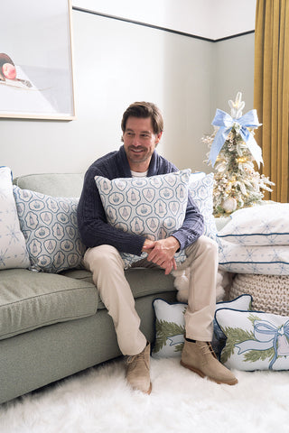 A man hugs the Silver Bells Pillow, featuring a festive winter design, while sitting on a green sofa. Twill-like pillows, a small Christmas tree with white ornaments and blue ribbon, and holiday decor add to the cozy living room atmosphere.