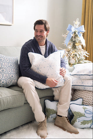 A man sits on a light green sofa, holding the Luxe Snowflakes Oversized Pillow. Behind him stands a small decorated Christmas tree with a blue bow and cushions nearby, including a blue pillow with winter patterns.