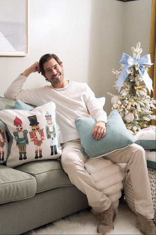 A smiling man in light clothes sits on a sofa with Sewing Down South holiday pillows, including the Pastel Nutcracker Pillow, next to a small Christmas tree adorned with white ornaments and a blue bow in a cozy, softly lit room.