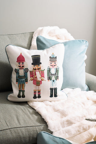 The Pastel Nutcracker Pillow, a white holiday accent featuring three illustrated nutcrackers, sits atop a light blue cushion and a white throw blanket on a light green armchair.