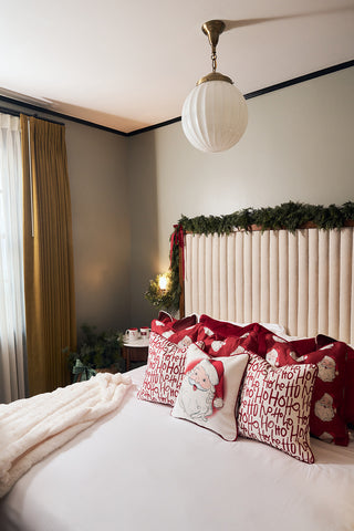 A cozy bedroom decorated for Christmas with a Vintage Stitch Santa Pillow and red holiday pillows featuring "Ho Ho Ho" text on the bed, a beige tufted headboard adorned with garland, and gold curtains by the window.