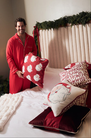 A smiling man in red pajamas holds a Vintage Repeating Santa Pillow in a festively decorated bedroom with holiday-themed pillows, greenery, and cozy bedding.