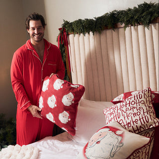 A man in red pajamas stands beside a bed holding a red pillow with white Santa faces. The bed is styled with multiple Christmas-themed pillows, including a white pillow with red 'Merry Christmas' text and another with a white Santa illustration.