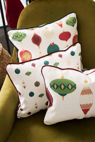 Three decorative accent pillows with colorful Christmas ornament patterns rest on a green armchair. Among them, a Hanging Ornaments Lumbar Pillow adds a touch of modern style with festive red, green, and gold baubles.