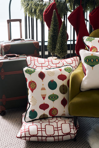 Two Classic Ornaments Pillows sit stacked by an olive-green chair. Red stockings and artificial greenery hang in the background, with green suitcases nearby.