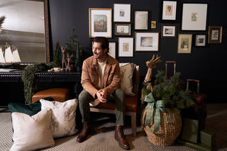 A man in a tan jacket and green pants sits on a bench in a festive room decorated with pine garlands, wrapped gifts, a Holiday Reindeer Pillow, a basket with greenery and ribbon, and framed art on a dark wall.