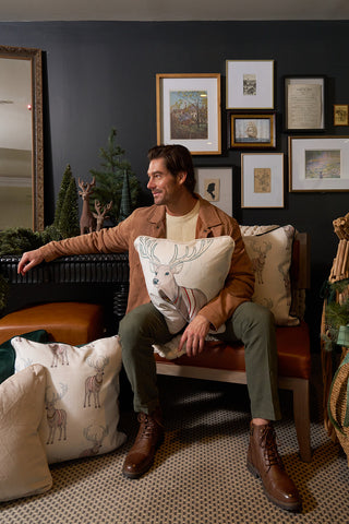 A man in a tan jacket and green pants sits on a bench holding the Holiday Reindeer Pillow, surrounded by festive decor and framed artwork on a dark wall, with other velvet deer-themed pillows nearby.