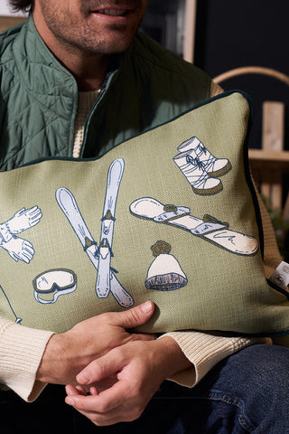 A man in a green vest and cream sweater holds a Winter Sports Gear Lumbar Pillow, an accent pillow featuring illustrations of skis, a snowboard, gloves, boots, goggles, and a knit hat.