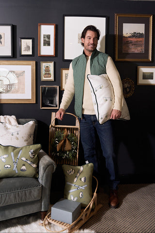 A man stands in a stylish living room with dark walls, holding the Ski Resort Pillow. Additional pillows and items rest in baskets nearby. Framed art above a green armchair enhances the room's chic apres-ski decor.