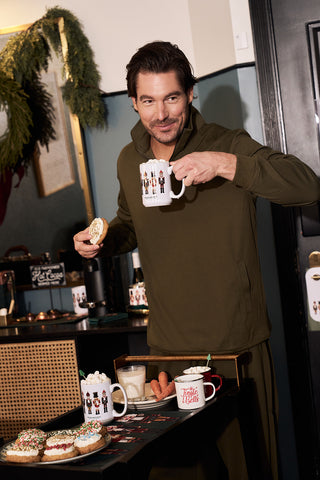A man in an olive green outfit smiles at the camera in a festive setting, holding a sprinkled donut and the Nutcracker Coffee Mug by a table with hot drinks and donuts—perfect for sharing as a holiday gift.