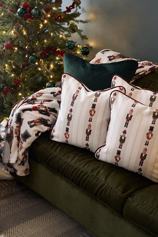A cozy green sofa featuring a Neutral Nutcrackers Pillow and festive blanket sits beside a Christmas tree decorated with lights and ornaments.