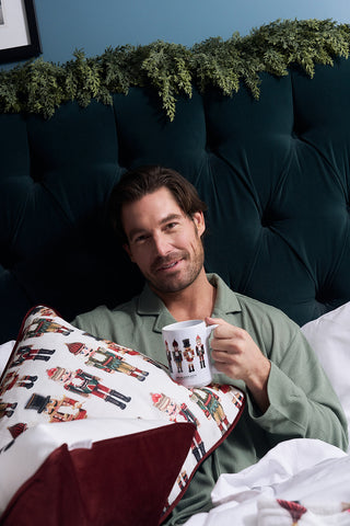 A man with brown hair sits in bed, holding the Nutcracker Coffee Mug. He wears green pajamas and leans against a dark green tufted headboard with greenery. A festive nutcracker-print pillow in front makes a perfect holiday gift.