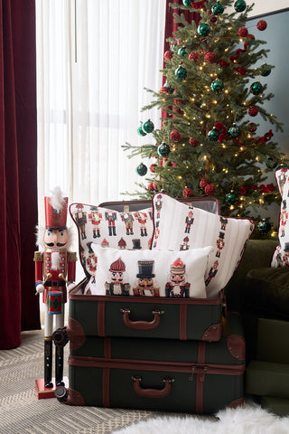 A decorated Christmas tree stands by a window with red curtains. Two stacked suitcases display Sewing Down South Nutcracker Holiday Lumbar Pillow, with a nutcracker figure beside them.
