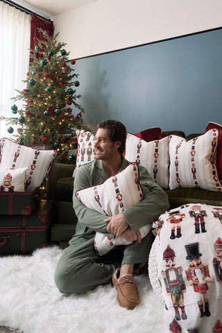 A man in green pajamas sits on a white rug, hugging a Neutral Nutcrackers Pillow. He is surrounded by holiday pillows with a Christmas tree decorated with ornaments and lights in the background.
