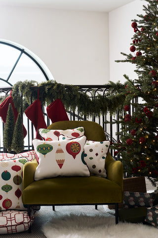 A green armchair with a Hanging Ornaments Lumbar Pillow rests beside a decorated Christmas tree. Red stockings hang on a garland-draped railing, while wrapped gifts with ornament patterns lie on the floor.