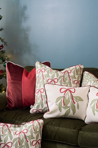 Several decorative pillows, including the Bows & Mistletoe Lumbar Pillow with a festive mistletoe design and solid red ones, are arranged on a dark green sofa. A decorated Christmas tree appears on the left, set against a light blue wall.