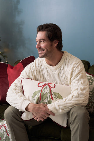 A man with brown hair and a beard, wearing a cream cable-knit sweater, sits on a green couch smiling and hugging the Bows & Mistletoe Lumbar Pillow, which features a festive mistletoe pattern and red ribbon. Holiday pillows are seen in the background.