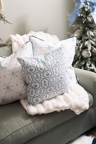 A cozy green couch with a Silver Bells Pillow, white snowflake-patterned pillows, and a throw blanket sits beside a decorated Christmas tree with snowy branches and a blue ribbon.