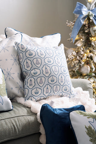 A cozy chair decorated with light blue and white throw pillows featuring snowflake and ornament patterns, next to a decorated Christmas tree with frosted branches and a large blue bow on top.