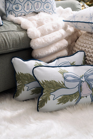 The Holiday Swag Extended Lumbar Pillow from Sewing Down South rests on a soft white rug beside a plush blanket and light green sofa, adding cozy, festive charm to the space.