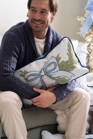 A smiling man in a navy sweater and beige pants sits on a sofa holding the Holiday Swag Extended Lumbar Pillow—a festive blue accent pillow with ribbon and greenery. A decorated Christmas tree is in the background.