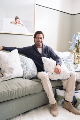 A man with brown hair smiles on a light couch adorned with the Luxe Snowflakes Oversized Pillow and white patterned pillows. To his right stands a Christmas tree, with framed art behind him, creating a cozy, festive setting.