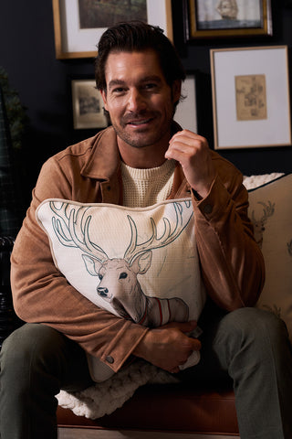 A man with brown hair and a beard, dressed in a tan jacket and cream sweater, sits on a couch holding the Holiday Reindeer Pillow. Framed art hangs on the dark wall behind him.
