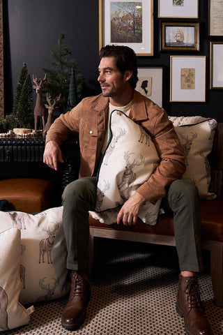 A man in a brown suede jacket and green pants sits on a couch, smiling and holding a Repeating Reindeer Pillow. He’s surrounded by deer-themed holiday décor, spruce green velvet pillows, miniature pine trees, and framed art on a dark wall.