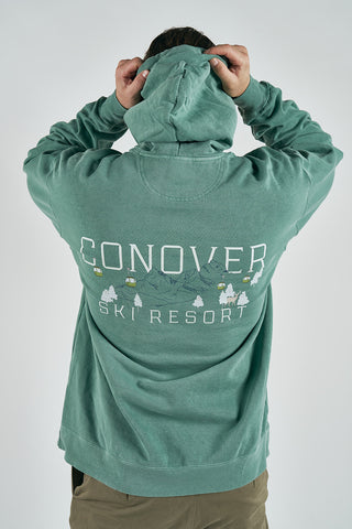 A person, facing away, pulls up the hood of a green Conover Ski Resort Hoodie with mountain graphics and cozy mountain-town style.