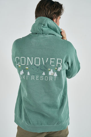 Wearing a green Conover Ski Resort Hoodie featuring illustrated mountains, trees, and cabins on the back, a person faces away with one hand near their chin, capturing the cozy charm of a mountain lodge.