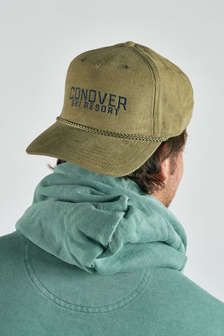 A person in a green hoodie and a Conover Ski Resort Embroidered Corduroy Hat, featuring blue lettering on khaki, brings mountain-town vibes to the look against a plain, light background.