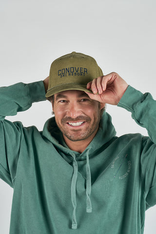 A smiling man in a green hoodie adjusts his Conover Ski Resort Embroidered Corduroy Hat, an olive-green cap with classic mountain-town style, against a plain light background.