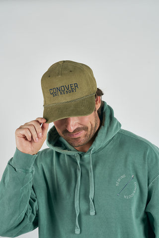 A man in a green hoodie tilts the brim of a Conover Ski Resort Embroidered Corduroy Hat, capturing classic mountain-town vibes against a plain light background.