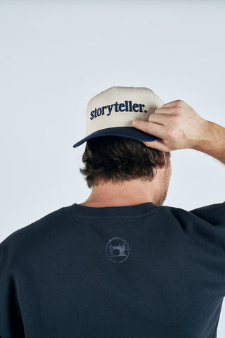 A person seen from behind adjusts a beige and navy Storyteller 3D Embroidered Hat, a 5-panel cap with an adjustable back, while wearing a dark t-shirt featuring a small circular logo on the back.