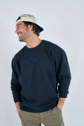 A man in a backward beige cap and Storyteller Embroidered Crewneck Sweatshirt smiles to the side with hands in his pockets, wearing khaki pants against a plain light background.