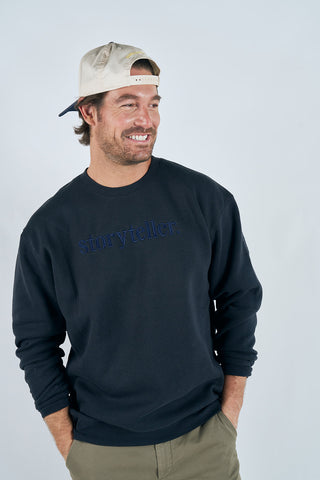 A man in a backwards beige cap, classic fit khaki pants, and a navy Storyteller Embroidered Crewneck Sweatshirt stands smiling with his hands in his pockets against a plain light background.