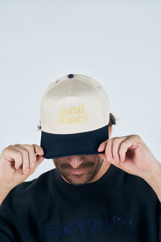 Wearing the Local Honey Embroidered Hat in yellow and a dark "storyteller." sweatshirt, a person holds the brim of their cap and looks downward.