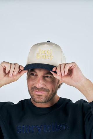 A bearded man smiles as he adjusts the Local Honey Embroidered Hat. He wears a navy sweatshirt with "storyteller." on the front, against a plain background that reflects his passion for beekeeping and supporting local products.