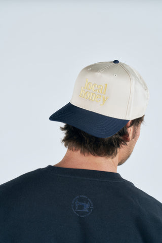 A person with short brown hair, seen from behind in a dark shirt, wears the Local Honey Embroidered Hat—a beige and navy cap with "Local Honey" stitched in yellow on the front, evoking a beekeeping vibe.