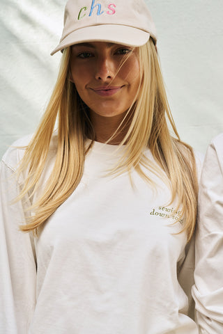 A young woman with long blonde hair wears the Rainbow Row Dad Cap—a beige hat with colorful “chs” letters—and a white “sew it down south” long-sleeve shirt, standing against a light background.
