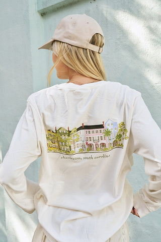 A person with long blonde hair, wearing a beige cap and a white Holiday Rainbow Row Long Sleeve Tee with a cityscape illustration on the back, stands with their back to the camera against a light wall.