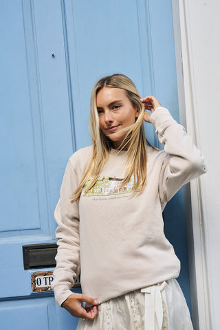 A young woman with long blonde hair stands in front of a light blue door, wearing the Holiday Rainbow Row Crewneck Sweatshirt and a white skirt. She touches her hair and gazes to the side with a relaxed expression.