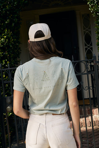 A woman in a light cap and pale green Christmas Tree Farm Tee stands facing a black iron gate in front of a house.