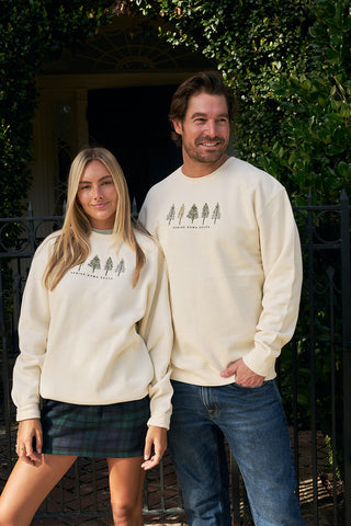 A man and woman stand outdoors by a black gate, both wearing matching Christmas Tree Farm Embroidered Crewnecks with a tree graphic. She pairs hers with a plaid skirt; he wears blue jeans. Green foliage frames the background.