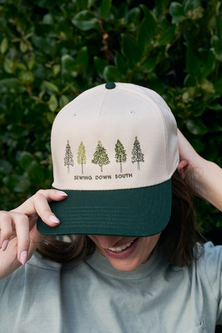 A person in the Christmas Tree Farm Embroidered Hat, featuring festive trees and “Sewing Down South,” smiles with hands on their head, standing before a backdrop of lush green leaves.