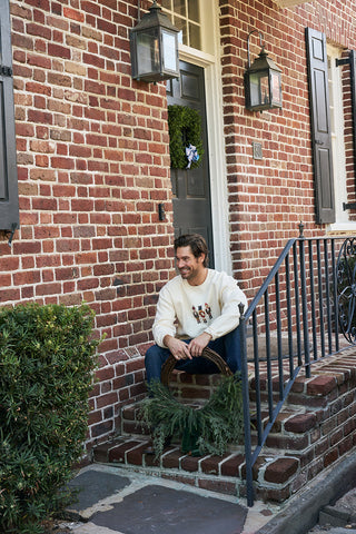 A man sits on brick steps outside a red brick house, holding a wreath with greenery. He wears a Nutcracker Crewneck Sweatshirt and jeans, with a green wreath hanging on the door behind him.