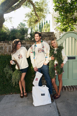 Three people stand outside by a green gate, smiling and holding greenery while wearing matching Nutcracker Crewneck Sweatshirts. The man in the center is wrapped in Christmas lights and a large white tote bag sits on the ground.