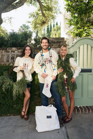 Three people smile in front of a garden gate, dressed for the holidays. The man in the center wears Christmas lights around his neck; one woman supports greenery, and another wears a Nutcracker Crewneck Sweatshirt.