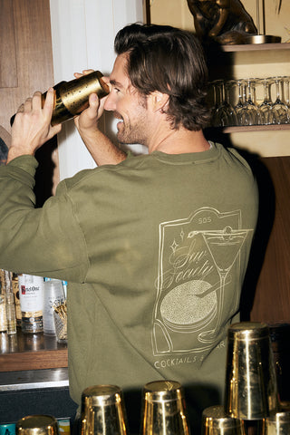 Wearing the olive green Cocktails & Caviar Crewneck Sweatshirt with a martini glass and citrus slice graphic, a man shakes a cocktail shaker behind the bar, surrounded by bottles and shakers.